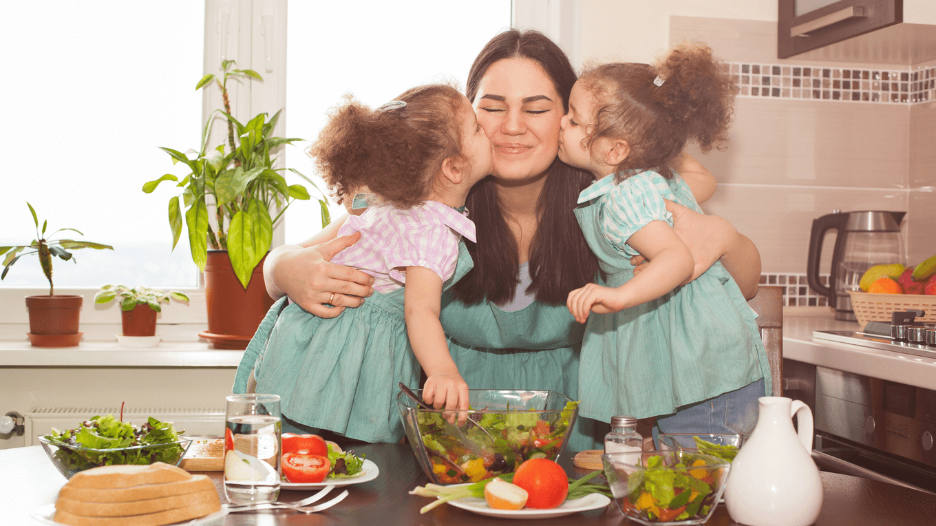 Усміхнена мама з двома маленькими дочками, які цілують її у щічки. На столі стоїть салат